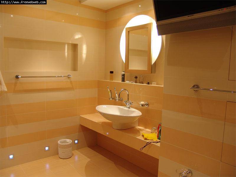 Jasna łazienka z poziomymi pasami płytek w odcieniach beżu i pomarańczy, okrągłe podświetlane lustro, biała umywalka nablatowa, wbudowane oświetlenie podłogowe i telewizor pod sufitem.